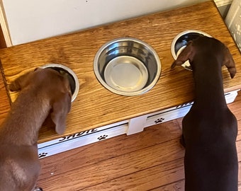May include: A raised wooden pet feeder station with a natural wood grain top and a white base featuring black paw print accents. The stand holds three circular stainless steel bowls, with two brown dachshunds eating from the outer bowls.