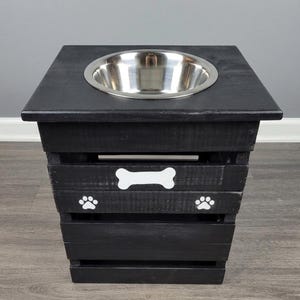 May include: A black wooden pet feeder stand with a square top and a single circular stainless steel bowl. The front of the stand features a white painted bone graphic centered above two white paw print graphics on the slatted wooden frame.