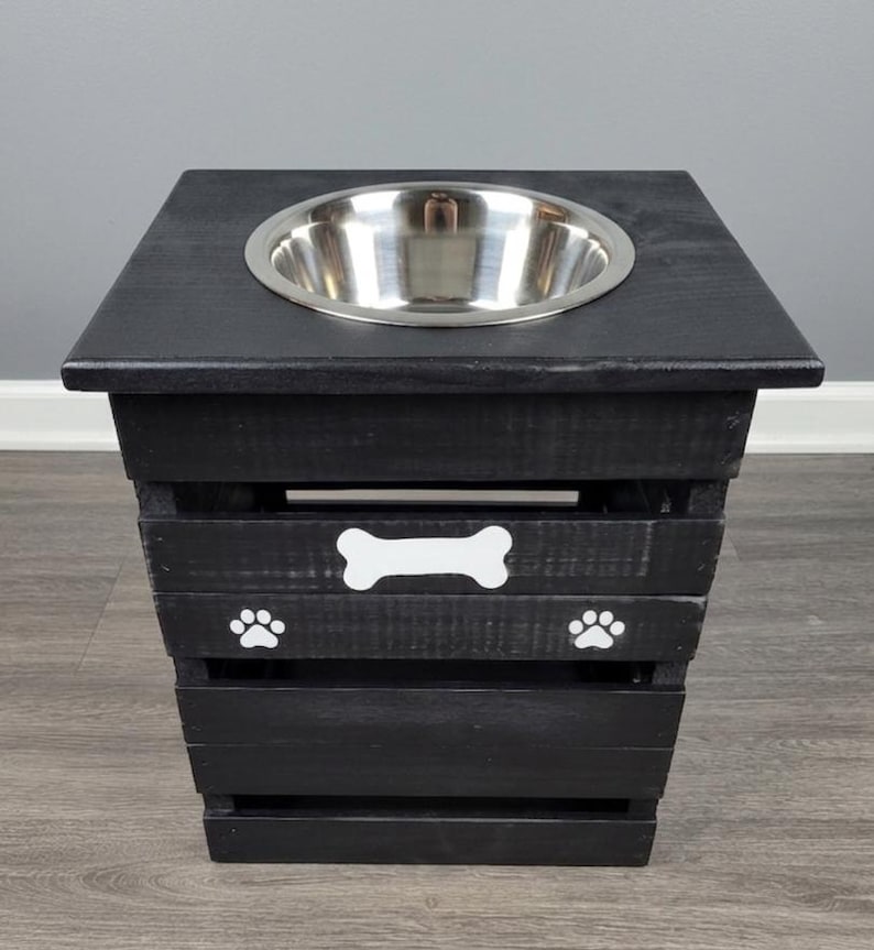 May include: A black wooden pet feeder stand with a square top and a single circular stainless steel bowl. The front of the stand features a white painted bone graphic centered above two white paw print graphics on the slatted wooden frame.