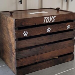 May include: A dark brown wooden storage crate with a lid featuring two small knobs. The front and top are decorated with white painted graphics including the word TOYS, a bone shape, and three paw prints. The crate has horizontal slats.