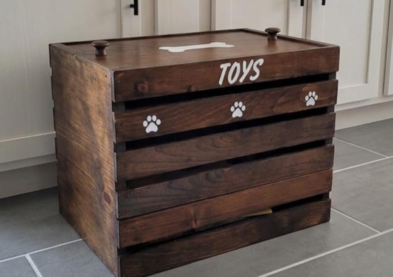 May include: A dark brown wooden storage crate with a lid featuring two small knobs. The front and top are decorated with white painted graphics including the word TOYS, a bone shape, and three paw prints. The crate has horizontal slats.