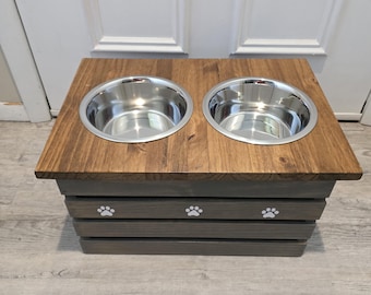 May include: A raised pet feeding station featuring a rectangular dark brown wooden top with two circular stainless steel bowls embedded side by side. The base is constructed from horizontal gray wooden slats and features three white paw print icons.