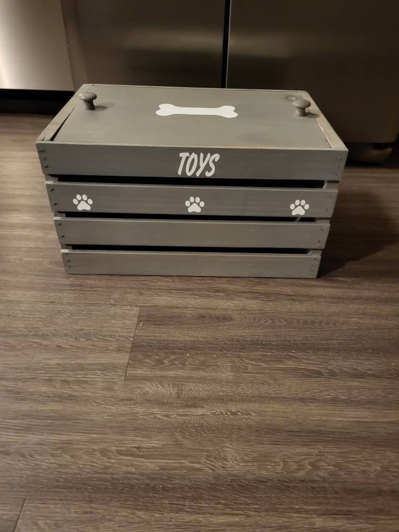 Puede incluir: Una caja de juguetes de madera gris con un dise&ntilde;o de hueso blanco y huellas de patas. La caja tiene una tapa y tres compartimentos. La tapa tiene dos pomos grises. La caja est&aacute; etiquetada como "TOYS".