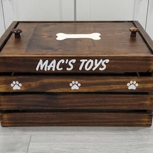 May include: A dark brown wooden crate with a lid featuring two small knobs. The front is labeled with the white text MAC'S TOYS and decorated with three white paw prints. A white bone shape is centered on the lid of this rectangular storage box.