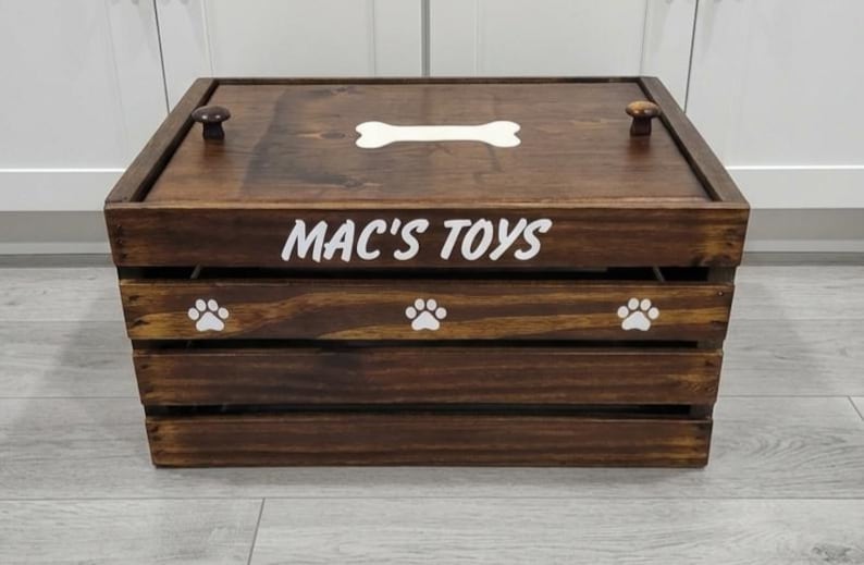 May include: A dark brown wooden crate with a lid featuring two small knobs. The front is labeled with the white text MAC'S TOYS and decorated with three white paw prints. A white bone shape is centered on the lid of this rectangular storage box.