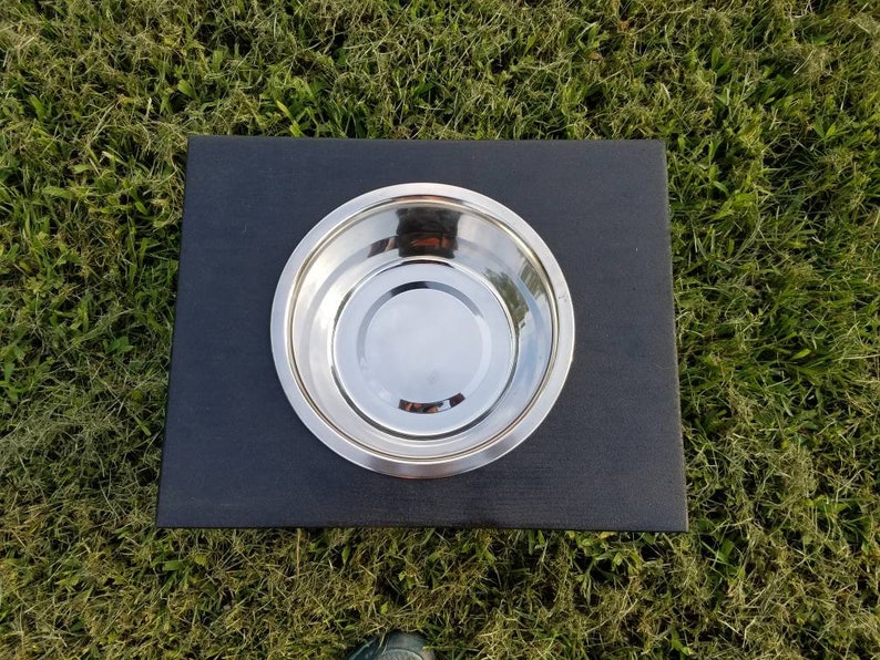 May include: A single circular stainless steel bowl set into a rectangular black base. The bowl features a polished, reflective metallic surface with a smooth, rounded rim, while the base is a solid, matte black platform with clean, straight edges.