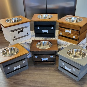 May include: Six elevated pet feeding stations made of wooden crates with a single stainless steel bowl on top. Each crate features a painted white bone icon and paw prints on the front. The colors include white, black, natural wood, and gray finishes.