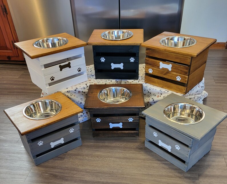 May include: Six elevated pet feeding stations made of wooden crates with a single stainless steel bowl on top. Each crate features a painted white bone icon and paw prints on the front. The colors include white, black, natural wood, and gray finishes.