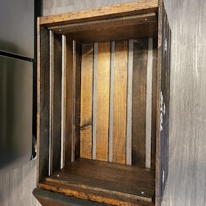 May include: A rectangular wooden crate with a dark brown stained finish. The interior base and sides are constructed from vertical wooden slats of varying brown tones. The word TOYS is printed in white capital letters on the exterior side panel.