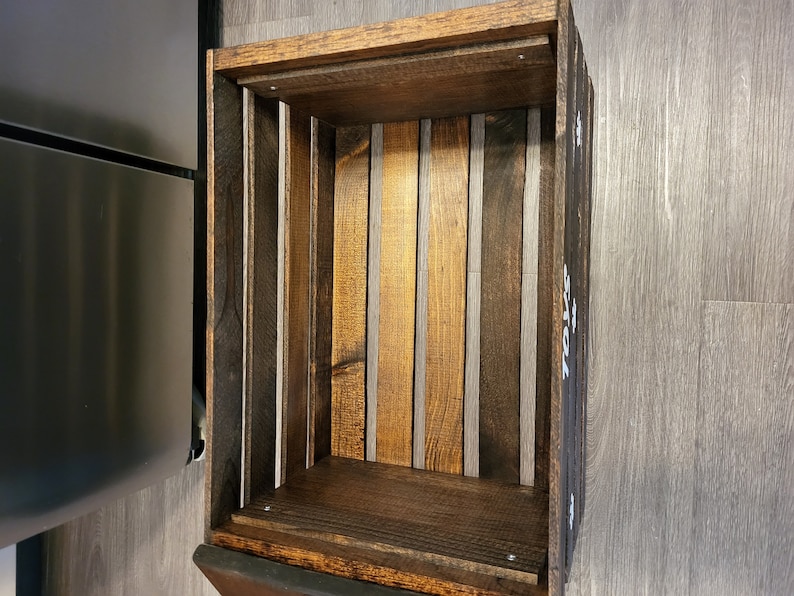 May include: A rectangular wooden crate with a dark brown stained finish. The interior base and sides are constructed from vertical wooden slats of varying brown tones. The word TOYS is printed in white capital letters on the exterior side panel.