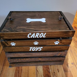 May include: A dark brown wooden storage crate with a lid featuring a white bone graphic. The front of the crate displays the white text CAROL and TOYS, separated by a slat with three white paw print graphics. The lid has two small round knobs.