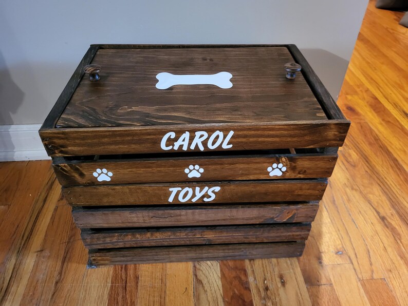May include: A dark brown wooden storage crate with a lid featuring a white bone graphic. The front of the crate displays the white text CAROL and TOYS, separated by a slat with three white paw print graphics. The lid has two small round knobs.