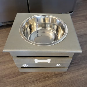 May include: A gray wooden dog bowl stand with a stainless steel bowl. The stand has a bone and paw print design.