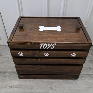 May include: A dark brown wooden storage crate with a lid featuring two small knobs. The top of the lid has a white bone graphic. The front of the crate displays the white text TOYS above three white paw print graphics on the top slat.