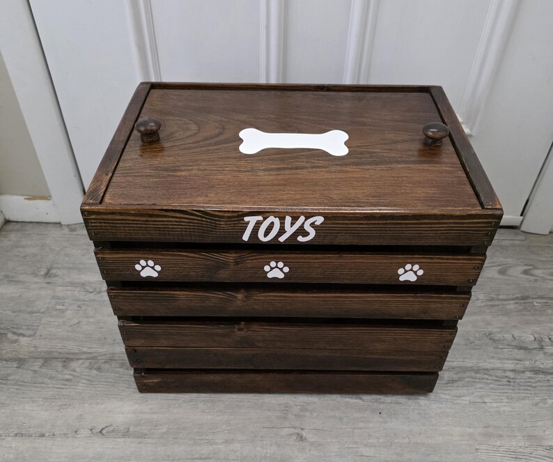 May include: A dark brown wooden storage crate with a lid featuring two small knobs. The top of the lid has a white bone graphic. The front of the crate displays the white text TOYS above three white paw print graphics on the top slat.