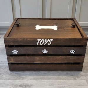 May include: A rectangular dark brown wooden crate with a lid featuring two small round knobs. The lid displays a white bone graphic, while the front panel features the white text TOYS above three white paw print graphics on the slats.