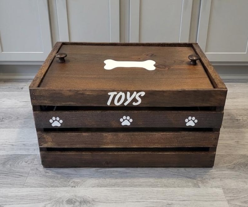 May include: A rectangular dark brown wooden crate with a lid featuring two small round knobs. The lid displays a white bone graphic, while the front panel features the white text TOYS above three white paw print graphics on the slats.