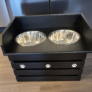 May include: A black wooden pet feeder station featuring two circular stainless steel bowls set into a raised platform. The front of the base is constructed with horizontal slats and is decorated with three white paw print icons centered horizontally.