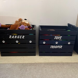 May include: Two black wooden slatted storage crates for pet items. The left crate features the white text RANGER and white paw prints. The right crate features the white text TOYS and TROOPER with white paw prints. Both crates are open.