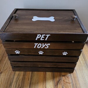 May include: A dark brown wooden crate with a lid featuring two small knobs and a white bone graphic. The front of the crate displays the white text PET TOYS and three white paw print graphics. The crate is constructed with horizontal slats.