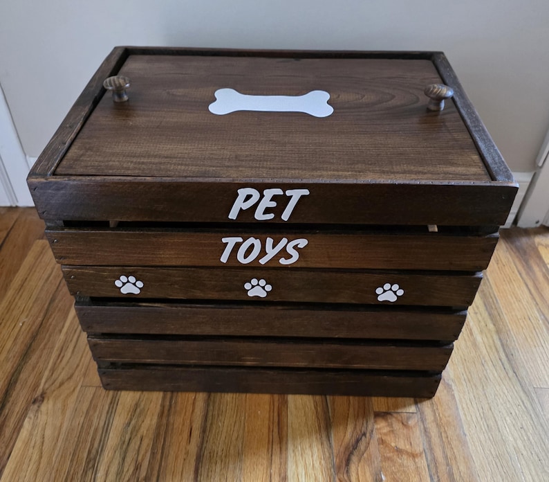 May include: A dark brown wooden crate with a lid featuring two small knobs and a white bone graphic. The front of the crate displays the white text PET TOYS and three white paw print graphics. The crate is constructed with horizontal slats.