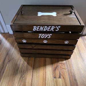 May include: A dark brown wooden crate with a lid featuring two small knobs. The top has a white bone graphic. The front panel displays the white text BENDER'S TOYS above three white paw print graphics. The crate is constructed with horizontal slats.