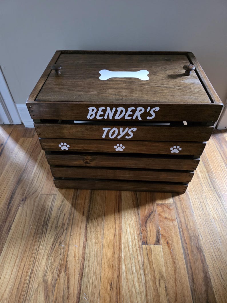 May include: A dark brown wooden crate with a lid featuring two small knobs. The top has a white bone graphic. The front panel displays the white text BENDER'S TOYS above three white paw print graphics. The crate is constructed with horizontal slats.