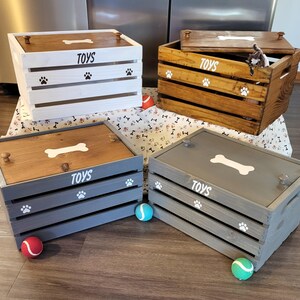 May include: Four rectangular wooden storage crates with lids, each featuring the word TOYS, a bone graphic, and paw print accents. The crates are finished in white, natural wood, and two shades of gray, with small knobs on the lids for easy opening.