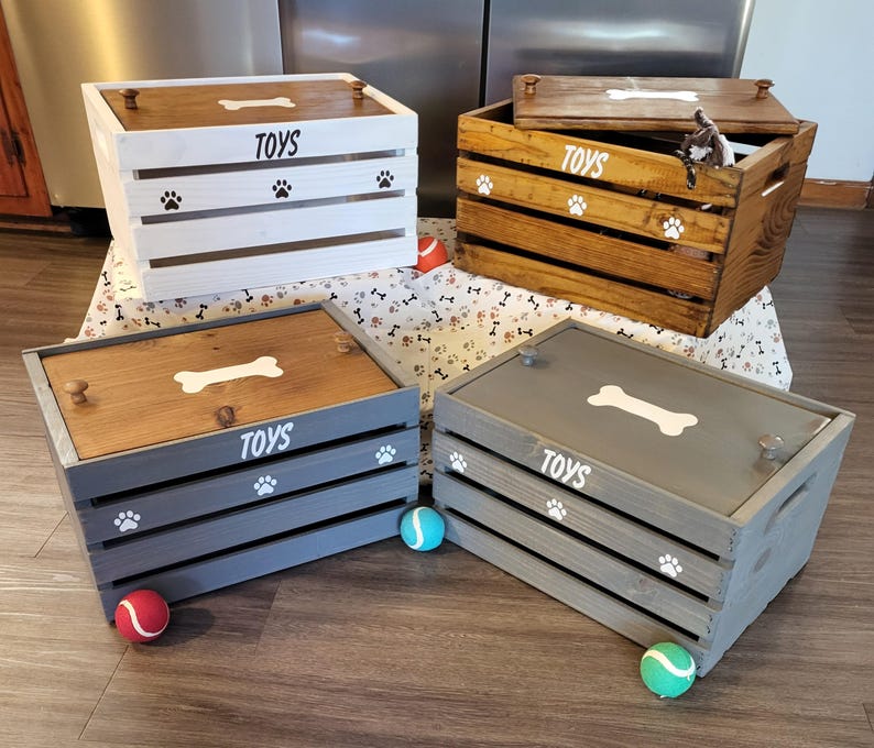 May include: Four rectangular wooden storage crates with lids, each featuring the word TOYS, a bone graphic, and paw print accents. The crates are finished in white, natural wood, and two shades of gray, with small knobs on the lids for easy opening.