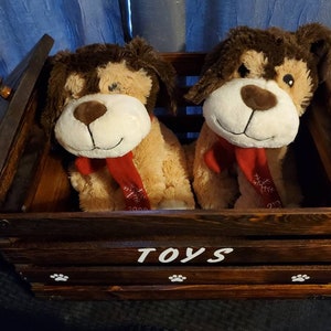 Puede incluir: Una caja de juguetes de madera con tapa con bisagras, pintada de marr&oacute;n con huellas de patas blancas y la palabra "TOYS". Dos juguetes de peluche de perro est&aacute;n dentro de la caja, uno marr&oacute;n y blanco y el otro tostado con una bufanda roja.