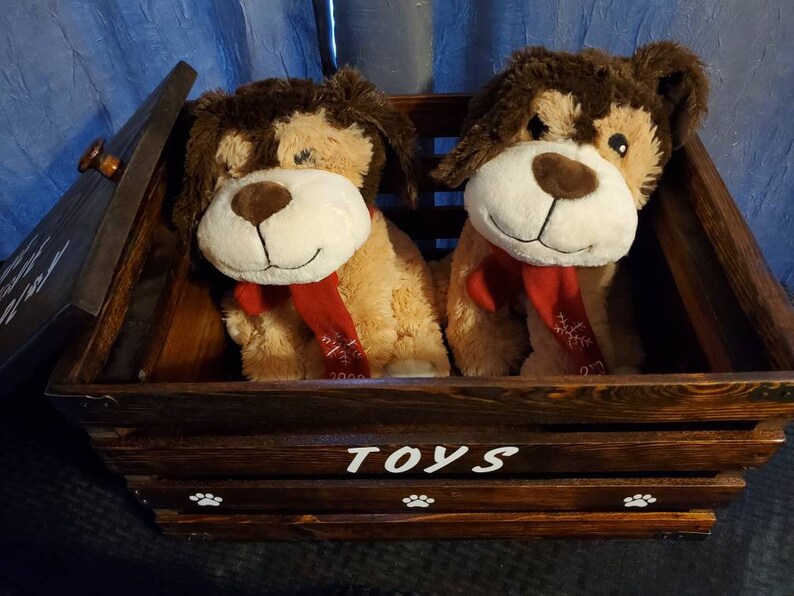 Puede incluir: Una caja de juguetes de madera con tapa con bisagras, pintada de marr&oacute;n con huellas de patas blancas y la palabra "TOYS". Dos juguetes de peluche de perro est&aacute;n dentro de la caja, uno marr&oacute;n y blanco y el otro tostado con una bufanda roja.