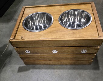 May include: A rectangular wooden pet feeder station featuring two circular stainless steel bowls set into the top surface. The front panel of the wooden crate-style base is decorated with three white paw print icons spaced evenly across the wood grain.