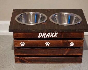 May include: A dark brown stained wooden pet feeder stand featuring two circular stainless steel bowls set into the top surface. The front panel displays the name DRAXX in white lettering above three white paw print icons centered on the slats.