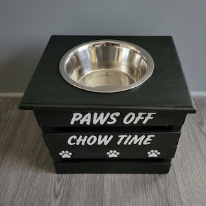 May include: A black wooden pet feeder stand with a single stainless steel bowl set into the top. The front features white text reading PAWS OFF and CHOW TIME, accompanied by three white paw print icons arranged horizontally along the bottom slat.
