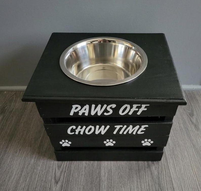 May include: A black wooden pet feeder stand with a single stainless steel bowl set into the top. The front features white text reading PAWS OFF and CHOW TIME, accompanied by three white paw print icons arranged horizontally along the bottom slat.