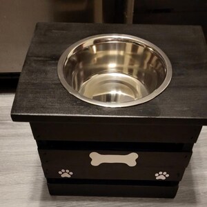 May include: A black wooden pet feeder stand with a square top and a single circular cutout holding a stainless steel bowl. The front panel features a white painted bone shape centered above two white painted paw prints, one on each side.