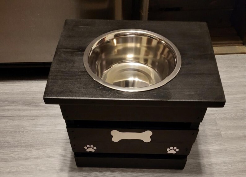 May include: A black wooden pet feeder stand with a square top and a single circular cutout holding a stainless steel bowl. The front panel features a white painted bone shape centered above two white painted paw prints, one on each side.