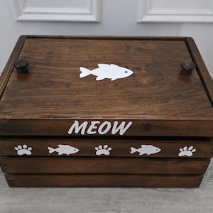 May include: A rectangular dark brown wooden crate with a lid featuring two small knobs. The lid displays a white fish graphic. The front panel is labeled with the white text MEOW and decorated with a repeating pattern of white fish and paw prints.
