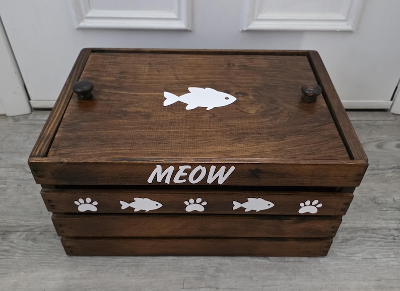 May include: A rectangular dark brown wooden crate with a lid featuring two small knobs. The lid displays a white fish graphic. The front panel is labeled with the white text MEOW and decorated with a repeating pattern of white fish and paw prints.