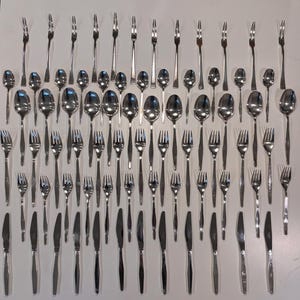 May include: A collection of stainless steel silverware, including forks, spoons, and knives, arranged on a white surface. The silverware is reflective, with a simple, modern design. The set includes various sizes of forks, spoons, and knives.