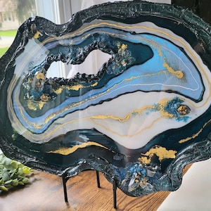 May include: A large, abstract resin art piece with a blue, white, and gold agate design. The resin is set on a black metal stand.