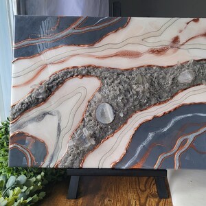 May include: Abstract painting on canvas with a white and gray background. The painting features a copper-colored design with a swirling pattern. The painting is on a wooden easel.