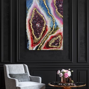 May include: Abstract wall art featuring a geode design with deep red, gold, and blue hues. The artwork has a textured appearance with gold accents. The piece is displayed on a black wall, above a gray armchair and a gold and black table.