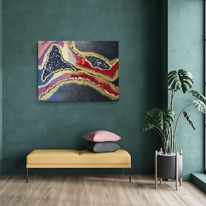 May include: Abstract art piece with a geode-like design. The artwork features a dark background with layers of red, gold, and blue, and a central area of dark blue crystals. The piece is mounted on a wall in a room with a yellow bench and a large plant.