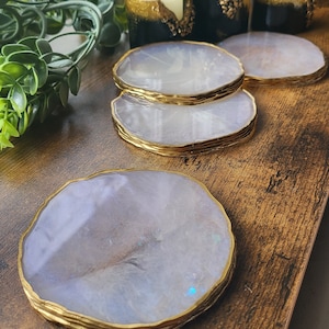 May include: Four white agate coasters with gold trim. The coasters are stacked on top of each other. The coasters are on a wooden surface.