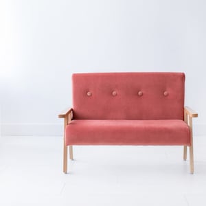 May include: A small, coral-colored loveseat with wooden armrests and legs. The seat back features four button details. The sofa is set against a white wall and floor, creating a minimalist aesthetic.