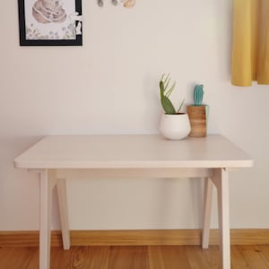 May include: A light-colored wooden table with four tapered legs. On the table sits a white rounded planter containing a green cactus and a cylindrical wooden container holding a small green artificial cactus. A framed illustration hangs above.