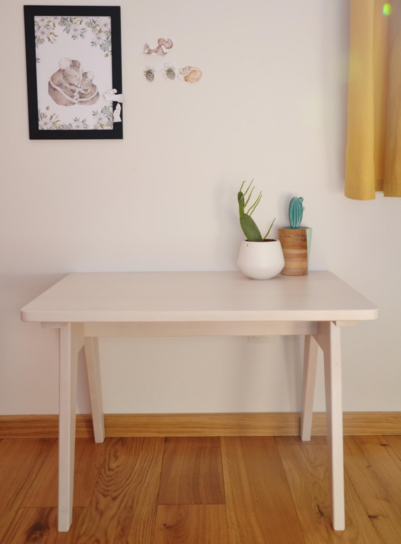 May include: A light-colored wooden table with four tapered legs. On the table sits a white rounded planter containing a green cactus and a cylindrical wooden container holding a small green artificial cactus. A framed illustration hangs above.