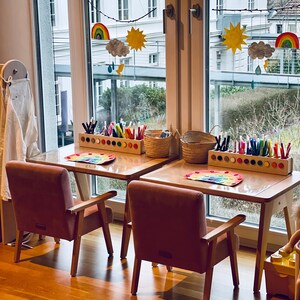 May include: Two light wooden children's desks with matching dusty pink upholstered armchairs. Each desk features a rectangular wooden organizer filled with colorful markers, a woven basket, and a paper rainbow craft placed on the tabletop.