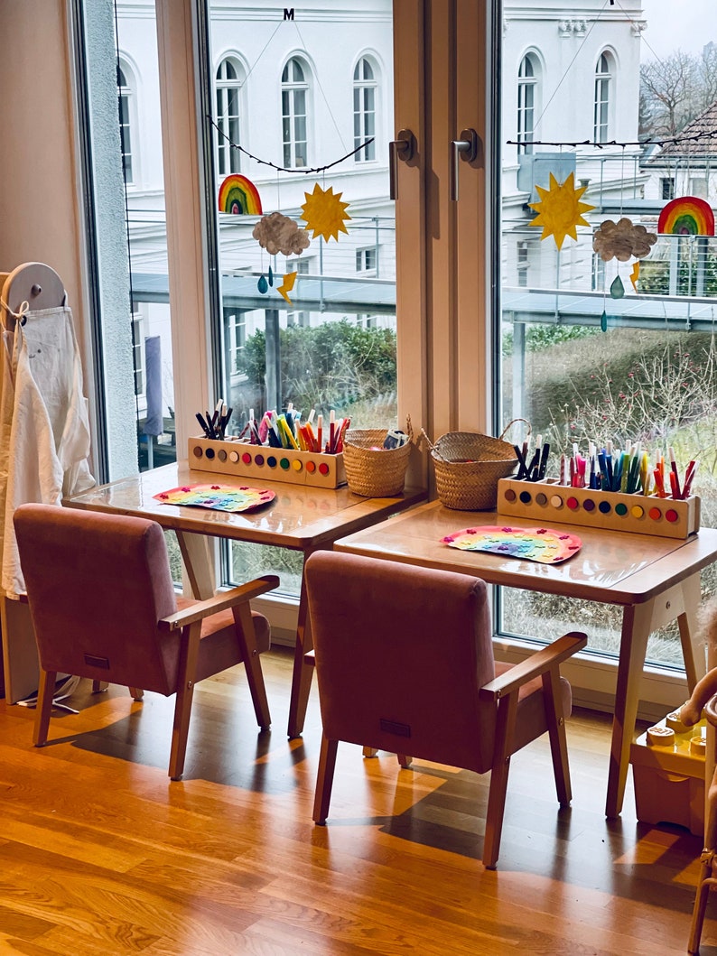 May include: Two light wooden children's desks with matching dusty pink upholstered armchairs. Each desk features a rectangular wooden organizer filled with colorful markers, a woven basket, and a paper rainbow craft placed on the tabletop.